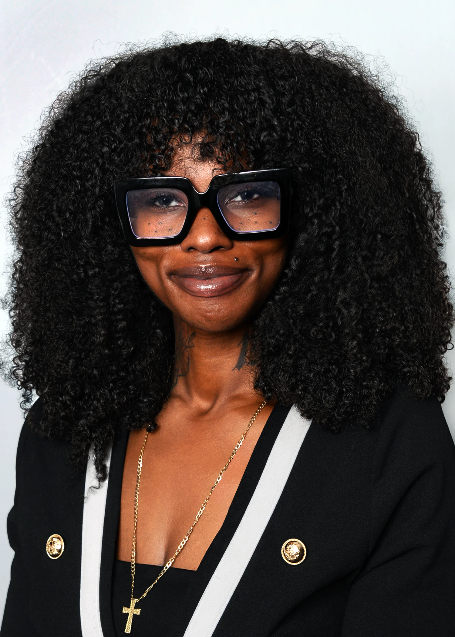 Portrait of a person with curly hair wearing large black square glasses and a cross necklace against a plain background.