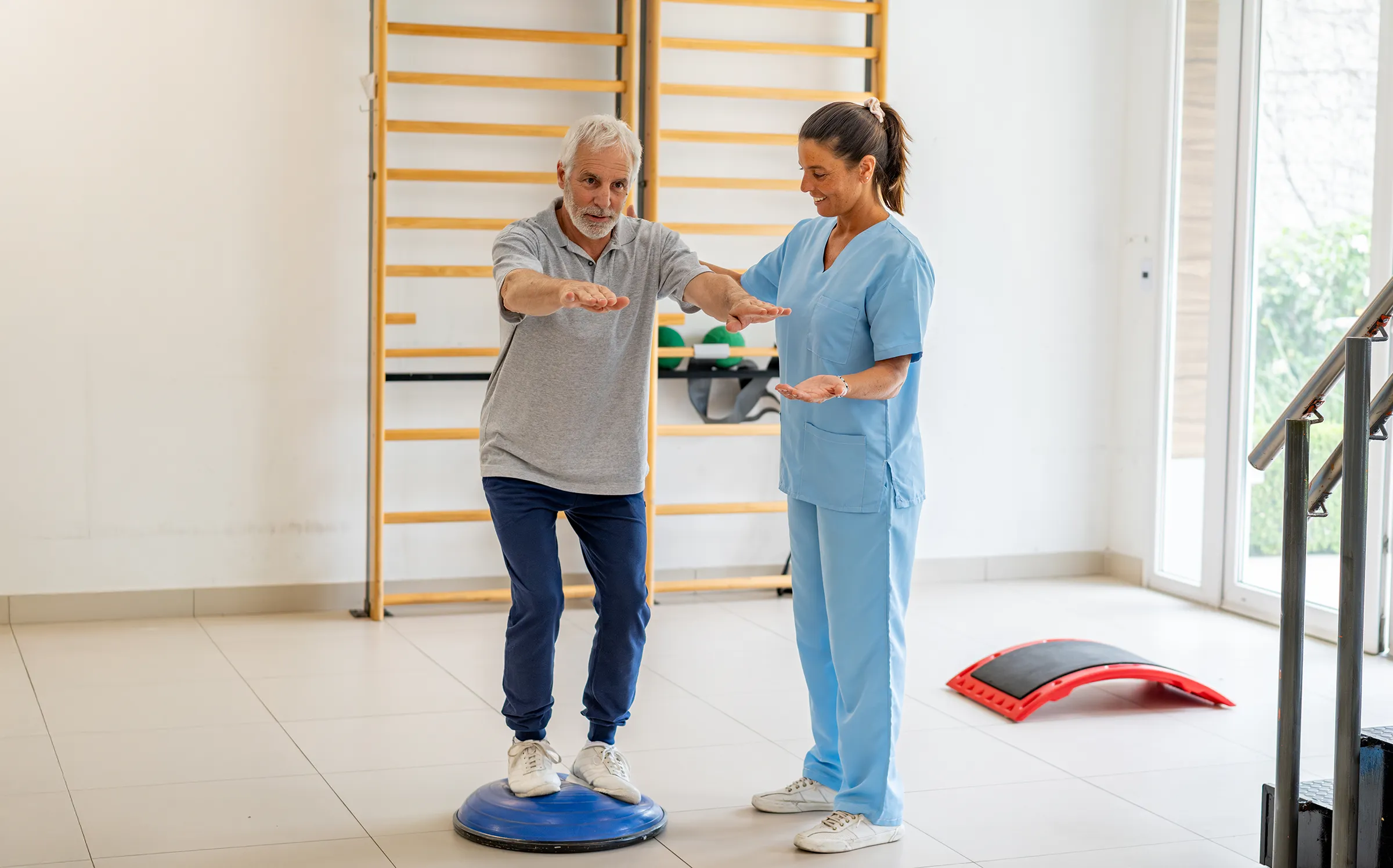 A caregiver assists a man standing on a balance trainer in a wellness room with therapy equipment and handrails.