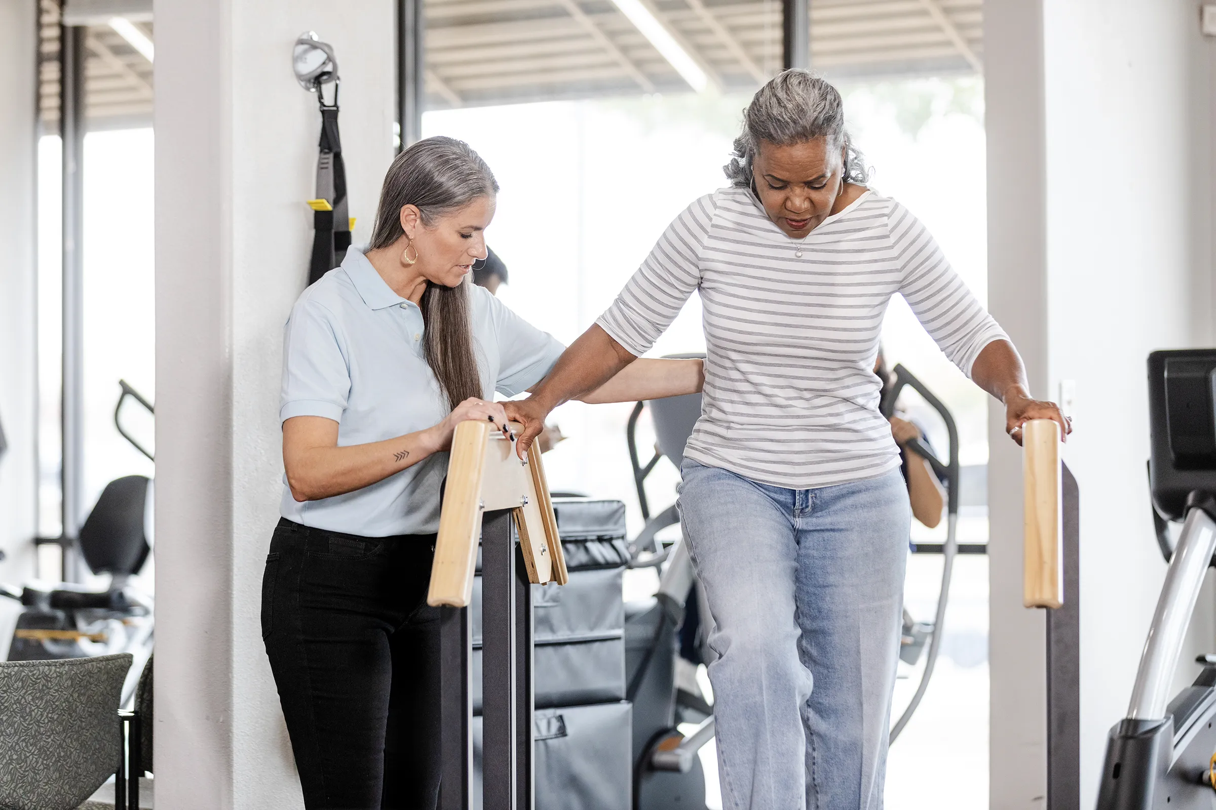 A senior woman practices balance exercises on parallel bars with assistance from a staff member in a fitness room at The Birches at New Hope.
